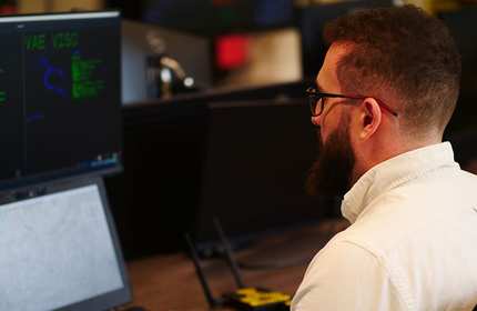 A man with a beard, wearing glasses, is sitting at a desk with multiple computer screens. He is focused on the monitors, which display various graphical data.