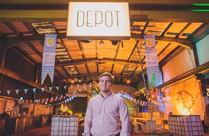 A person in a pink shirt stands under a sign that reads DEPOT inside a large, colorfully lit indoor space. The area is decorated with banners and string lights, creating a festive atmosphere.