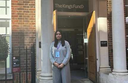A young woman wearing a lanyard and ID badge stands outside the entrance to a building. Above the entrance is a sign that reads The King's Fund.