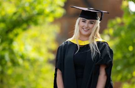 A young woman wearing a graduation cap and gown stands smiling. In the background is green foliage.