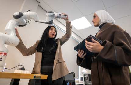 A young woman holding a tablet looks on as another woman manipulates a robotic arm.