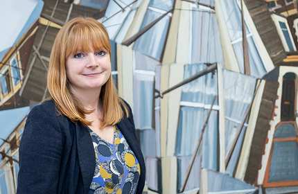 A woman with red hair stands in front of an abstract, geometric painting featuring overlapping architectural elements. She wears a patterned top under a dark jacket and smiles gently at the camera.