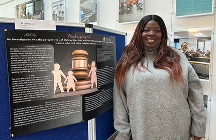 A person stands smiling next to a display board with a research poster titled An investigation into the perspectives of child protection workers on domestic abuse. The poster features text and an image of a gavel with figures of a child and parents.