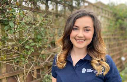 A smiling person with wavy hair wearing a navy shirt with a logo stands outdoors in front of a wooden lattice covered with vines.