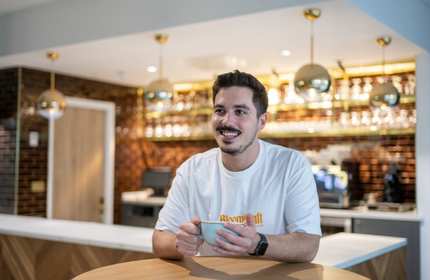A young man sits at a round table holding a coffee cup in his hands. In the background is a bar area.