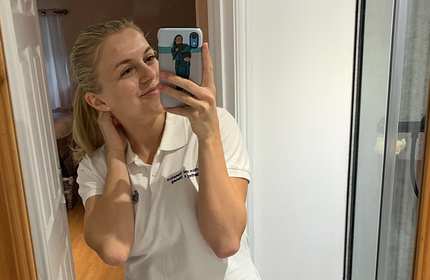 A person with blonde hair takes a mirror selfie, smiling and touching their neck. They are wearing a white polo shirt with embroidered text. The reflection of a room is visible in the mirror.
