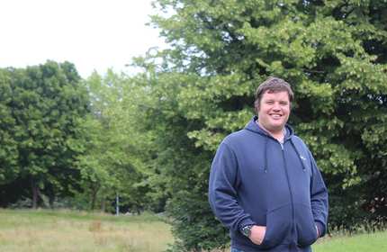 A person wearing a navy blue hoodie stands smiling with hands in pockets in a grassy field. Lush green trees fill the background under a cloudy sky.