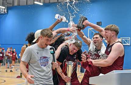 A person stands on a basketball court holding a microphone, while a group of players celebrate in the background.