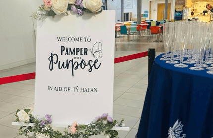 A sign with floral decorations reads Welcome to Pamper for a Purpose in aid of T≈∑ Hafan. Beside it, a table holds numerous empty champagne flutes, and colorful chairs are visible in the background.