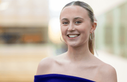 A young woman with hoop earrings and a nose ring smiles while wearing an off-the-shoulder blue top. The background is blurred, highlighting her cheerful expression.