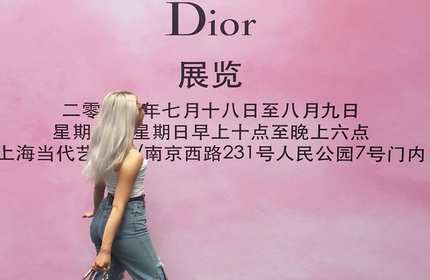 A person with long blonde hair, wearing a white top and jeans, stands in front of a pink wall with black text featuring the word Dior and information in Chinese characters.