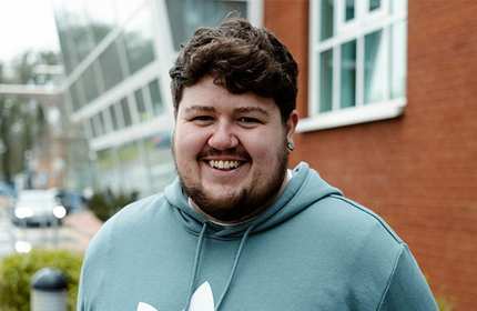 A person with short, curly hair and a beard is smiling and wearing a green hoodie with a white logo. They are standing outdoors near a modern building with glass windows and a brick wall. Its a cloudy day.