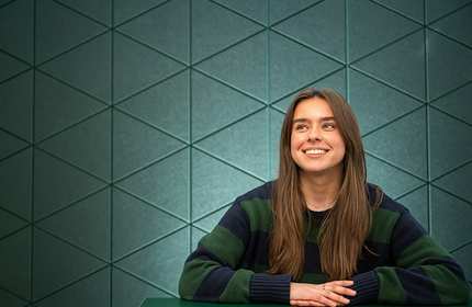 A person with long brown hair sits at a table, smiling and looking upward. They are wearing a dark green and black striped sweater. The background is a geometric patterned wall in shades of green.