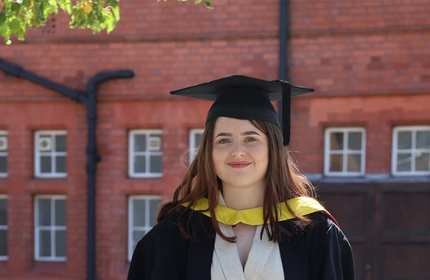 A graduate wearing a black cap and gown with a yellow stole stands smiling in front of a brick building with windows. Bright green leaves frame the upper left corner of the image.
