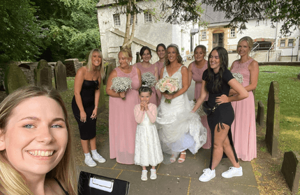 A bride, dressed in white and holding a bouquet, poses with bridesmaids in pink dresses and a young girl in a white dress. They stand in front of an old building and gravestones, with a smiling woman taking a selfie in the foreground.