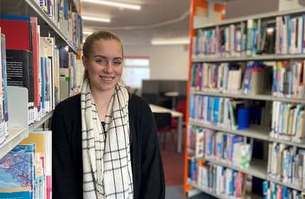 A person wearing a black sweater and a checkered scarf smiles while standing next to bookshelves in a library. The background features more shelves filled with books and a table with chairs.