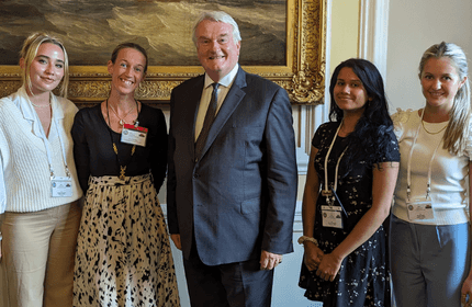 A group of five people, four women and one man, stand smiling in a formal setting. They are wearing business attire and conference badges. A large painting hangs on the wall behind them.
