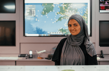 A woman wearing a gray hijab stands in front of a display showing a world map with various points marked. She is smiling, and there are computers and office equipment around her.