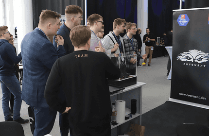 A group of people stands near a table with a computer setup, engaging in discussion. They are in an indoor event space with banners displaying the word Covenant and accolades like Great Place to Work in the background.