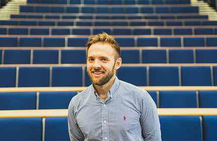 A man with a beard and short hair, wearing a checkered shirt, is smiling while standing in a large lecture hall with rows of empty blue seats.
