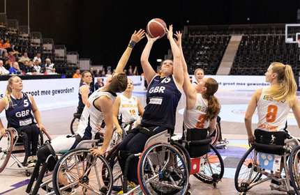 A wheelchair basketball game in progress between team GBR and team Netherlands. A player from team GBR is attempting a shot while being defended by team Netherlands players. Other players and the audience are visible in the background.