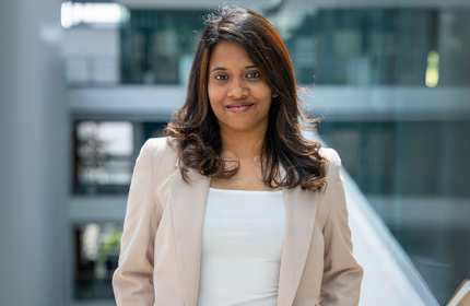 A person in white shirt and beige jacket stands in a brightly-lit atrium space.