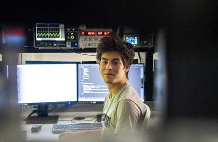 A young person in a t-shirt sits at a desk in front of two computer monitors. Above the monitors is an oscilloscope and several other electronic devices.