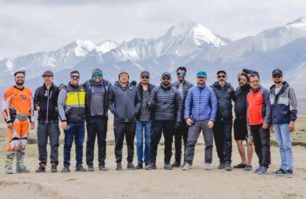 A large group of people stand in a row. In the background is a mountain range.