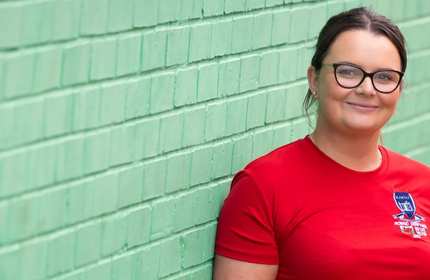 A smiling person leans against a green painted brick wall.
