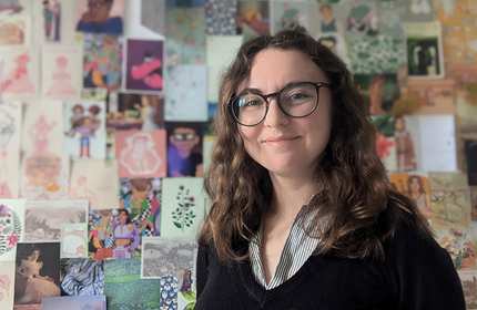 A person stands smiling in front of a wall covered with postcards and prints.