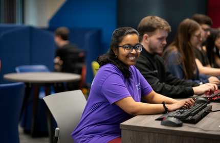 A person types at the keyboard of a computer workstation. Behind them are other people also using workstations.