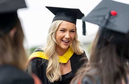 A young woman wearing a graduation cap and gown smiles brightly during a graduation ceremony. She stands among fellow graduates, celebrating their achievements. The background is slightly blurred, highlighting the sense of joy and accomplishment.