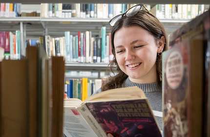 A young woman with glasses on her head smiles while reading a book in a library. Shelves filled with various books surround her.