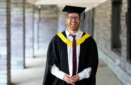 A person wearing a graduation cap and gown stands in an outdoor covered walkway.