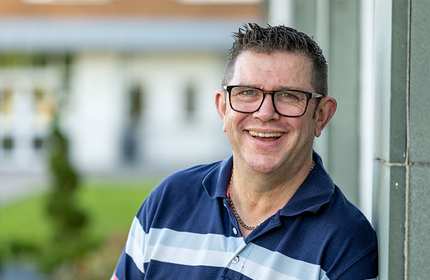 A man wearing glasses and a striped polo shirt smiles while leaning against a green wall outdoors. The background features blurred greenery and buildings.