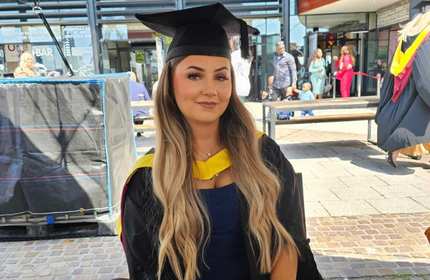 A young woman wearing a graduation cap and gown sits smiling. In the background are benches and groups of people walking by.