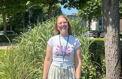 A smiling woman with long light red hair wearing glasses, a white “Riviera” t-shirt with a pink graphic, and a blue lanyard stands outdoors in front of tall grasses and trees on a sunny day.