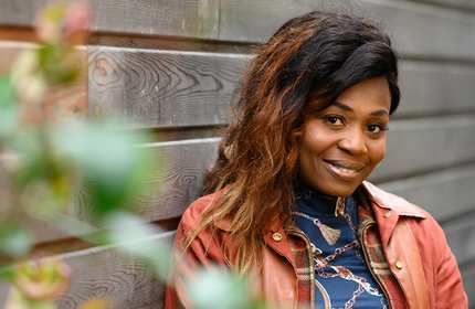 A woman with long hair smiles at the camera while leaning against a wooden wall. Shes wearing a brown leather jacket and a patterned shirt. In the foreground, out-of-focus green leaves add a touch of nature to the scene.
