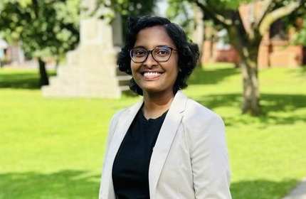 A woman wearing a white suit jacket and black shirt stands on a path with a grass field a trees in the background