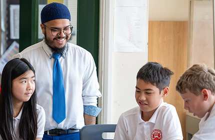 Teacher wearing shirt and tie stands behind table of three pupils