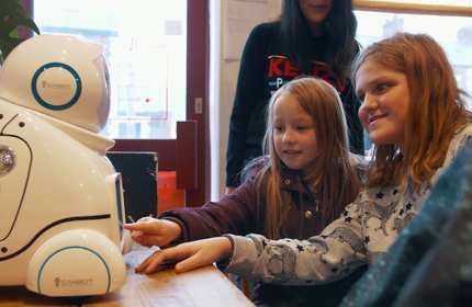 Two young girls interact with a screen on the centre of a small humanoid robot