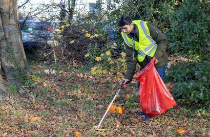 Young adult in high visibility jacket uses a litter picker to collect rubbish in a large red bin bag
