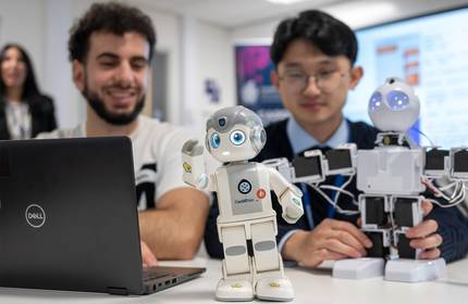 Two men interacting with robots in a tech setting. One is using a laptop, while two humanoid robots are on the table. The foreground robot is small and white with a delighted expression. The background has monitors and a blurred person.