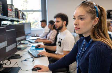 A group of people sit alongside each other at a long desk. Each of them is using a computer workstation.
