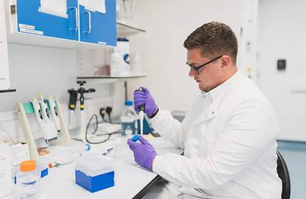 A person in a lab coat uses a pipette to dispense a sample into a container.