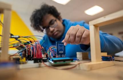 A person in a blue hoodie works with electronic components and circuit boards, placing a small blue board among connecting wires on a table. The background is blurred and the lighting is bright.