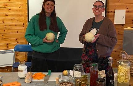 Two women holding cabbages stand behind a table full of pickling liquids and vegetables