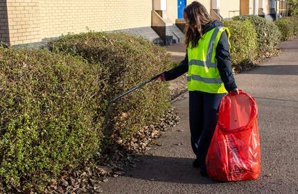 A woman in a high-visibility jacket reaches for rubbish in a bush with a litter picker