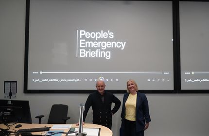 Two individuals stand at the front of a lecture theatre in front of a large presentation screen that reads 'People's Emergency Briefing'