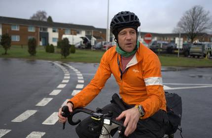 A man in high visibility clothing and a bicycle helmet rides a bicycle along campus roads